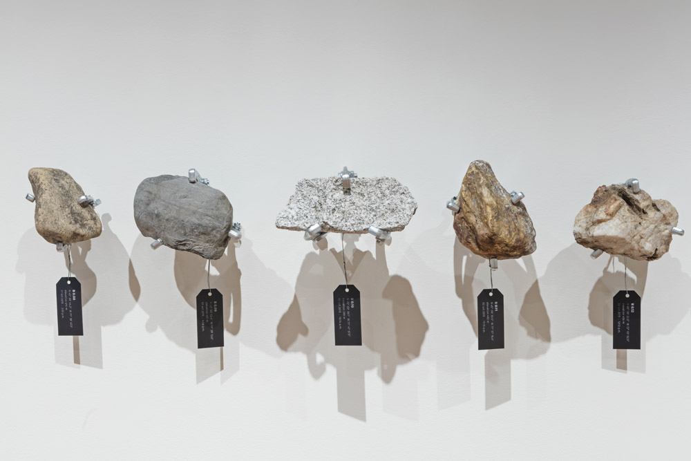rocks suspended in front of a wall, with black tags hanging from them with GPS co-ordinates